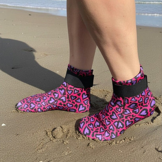 Person wearing pink and purple heart-patterned neoprene swimming socks, standing at the water's edge on sand