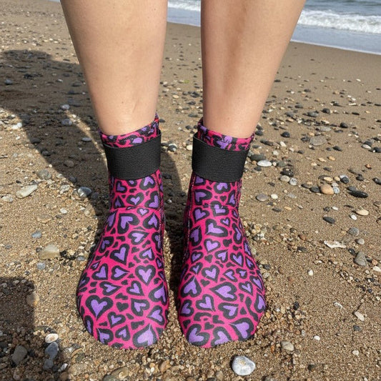 Person wearing pink and purple heart-patterned neoprene swimming socks, standing on a sandy beach