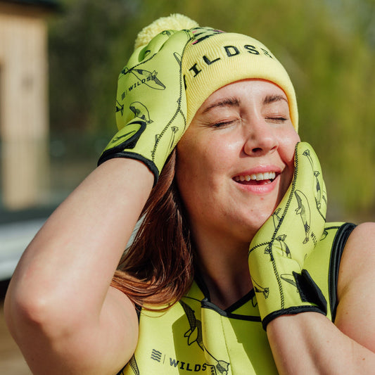 Person wearing yellow leopard print hat and neoprene swimming gear by water.