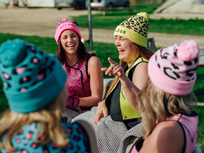 Four women wearing colorful beanies sitting outdoors on a grassy area.