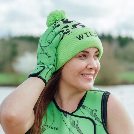 Person wearing green leopard print hat and neoprene swimming gear by water.